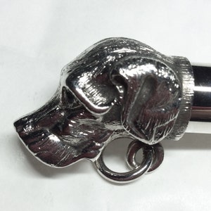May include: Silver whistle with a detailed dog head design. The whistle has a ring attached to the side.