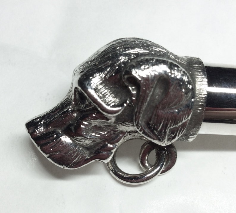 May include: Silver whistle with a detailed dog head design. The whistle has a ring attached to the side.