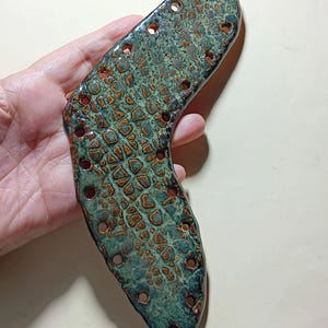 May include: A ceramic tile with a green and brown glaze and a crocodile skin pattern. The tile has multiple holes around the edges.