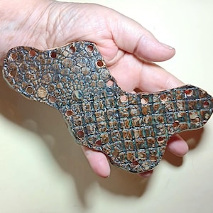 May include: A ceramic tile with a textured surface resembling crocodile skin. The tile is brown, green, and black with a pattern of small circles and squares. It has multiple holes around the edges.