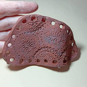 May include: A terracotta clay tile with a decorative pattern of leaves and swirls. The tile has multiple holes around the edge.