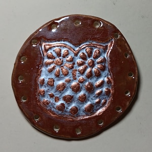May include: A brown ceramic owl ornament with white painted details. The owl has a round body and large eyes. The ornament has holes around the edge.