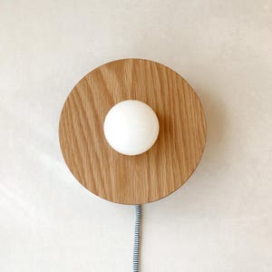 May include: A modern wall sconce featuring a round, light-colored wood base with a white globe light in the center. A blue and white striped cord hangs below. The fixture is mounted on a white wall.