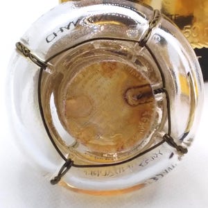 May include: A clear glass bottle with a cork and wire cage closure. The bottle has text printed on the glass and a gold-colored label. The cork is visible through the glass, and the wire cage is wrapped around the neck of the bottle.