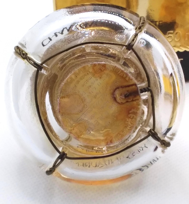 May include: A clear glass bottle with a cork and wire cage closure. The bottle has text printed on the glass and a gold-colored label. The cork is visible through the glass, and the wire cage is wrapped around the neck of the bottle.