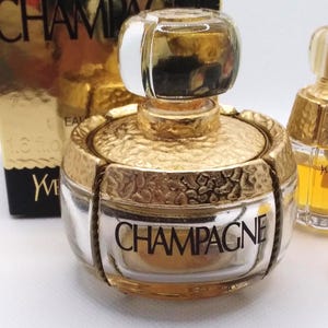 May include: A gold-toned perfume bottle with a clear glass body and a textured gold cap. The word "CHAMPAGNE" is printed on the front of the bottle. The bottle is part of a fragrance collection.