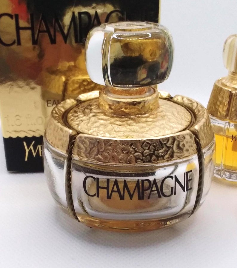 May include: A gold-toned perfume bottle with a clear glass body and a textured gold cap. The word "CHAMPAGNE" is printed on the front of the bottle. The bottle is part of a fragrance collection.