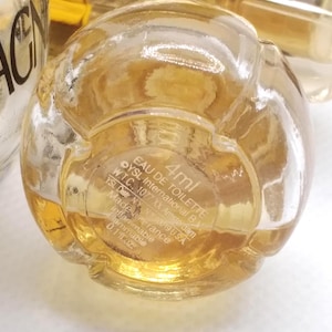 May include: A clear, spherical perfume bottle with a golden liquid inside. The bottom of the bottle features printed text including "EAU DE TOILETTE" and "4ml". The bottle is labeled "YSL International B.V." and "0.1 fl.oz."