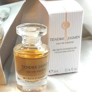 Könnte beinhalten: Ein kleiner Parfümflakon mit einem transparenten Glaskörper und einer goldfarbenen Flüssigkeit. Auf der Flasche steht "Tendre Jasmin Eau de Parfum" und wird von einer passenden Schachtel begleitet. Auf der Schachtel und der Flasche steht "Secrets d'Essences Yves Rocher Parfums". Die Flasche ist 5 ml.