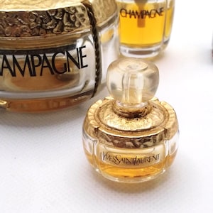 May include: Miniature perfume bottles with gold-colored accents. One bottle is labeled "Yves Saint Laurent" in black text. Other bottles are visible, including one labeled "CHAMPAGNE". The bottles are filled with a golden liquid.