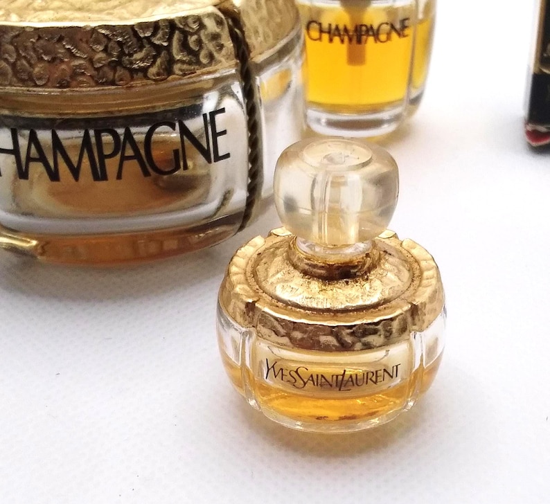 May include: Miniature perfume bottles with gold-colored accents. One bottle is labeled "Yves Saint Laurent" in black text. Other bottles are visible, including one labeled "CHAMPAGNE". The bottles are filled with a golden liquid.