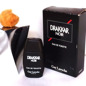 Peut inclure: Un flacon et une boîte noirs de Drakkar Noir Eau de Toilette. Le flacon est de petite forme rectangulaire avec le logo Drakkar Noir en blanc. La boîte est noire avec le logo et le texte Drakkar Noir en blanc et rouge. La boîte indique 5 ml et .17 fl oz.