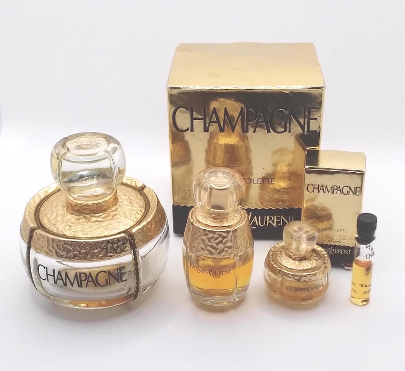 May include: A collection of gold-toned perfume bottles and boxes. The bottles have clear glass stoppers and are labeled "Champagne" and "Yves Saint Laurent". The boxes are gold with the word "Champagne" printed on them.