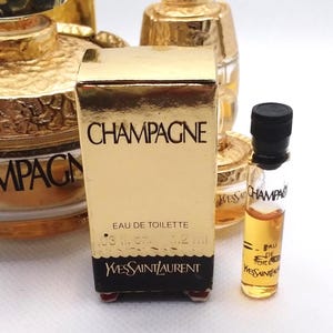 May include: A collection of miniature perfume bottles, including a gold-colored box labeled "CHAMPAGNE" by Yves Saint Laurent. The box contains "EAU DE TOILETTE" and a small glass vial with a black cap, also labeled "CHAMPAGNE."