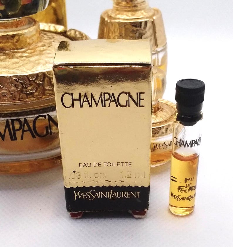 May include: A collection of miniature perfume bottles, including a gold-colored box labeled "CHAMPAGNE" by Yves Saint Laurent. The box contains "EAU DE TOILETTE" and a small glass vial with a black cap, also labeled "CHAMPAGNE."