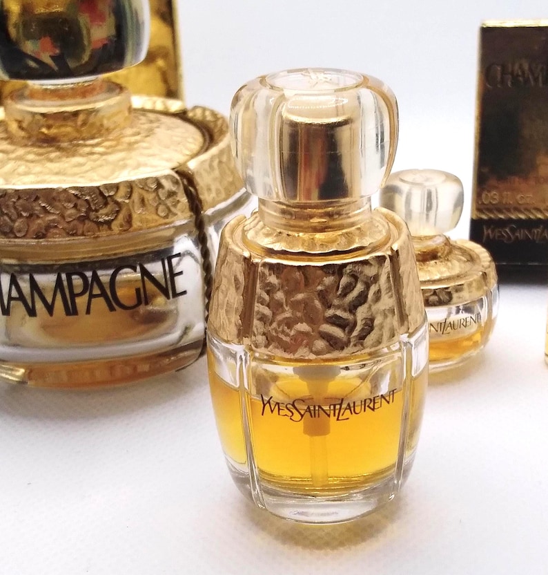May include: Several miniature perfume bottles with gold accents and clear glass. The bottles have a textured gold top and a clear glass base. The bottles are labeled with the brand name "Yves Saint Laurent" and the word "Champagne".