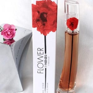 Peut inclure: Un coffret parfum avec une bouteille et une boîte. La boîte blanche présente un grand coquelicot rouge et le texte "FLOWER BY KENZO L'ABSOLUE". Le flacon de parfum est transparent avec une fleur rouge à l'intérieur. Une petite rose rose est à gauche.