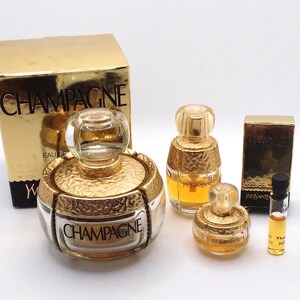 May include: A collection of gold-toned perfume bottles and boxes. The bottles vary in size and shape, with the word "CHAMPAGNE" visible on one. The boxes are gold with black text. A small vial of perfume is also present.