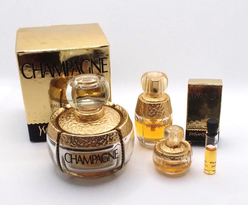 May include: A collection of gold-toned perfume bottles and boxes. The bottles vary in size and shape, with the word "CHAMPAGNE" visible on one. The boxes are gold with black text. A small vial of perfume is also present.