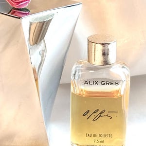 May include: A small glass bottle of Alix Grès Eau de Toilette with a gold-toned cap, containing 7.5 ml of amber liquid. The label features the text Alix Grès, a signature, Eau de Toilette, 7.5 ml, and Grès Paris, next to a silver-toned object with a pink rose.