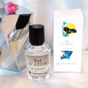Könnte beinhalten: Ein klarer Parfümflakon mit schwarzem Verschluss, beschriftet mit "Sel D'Azur Eau de Parfum" von Yves Rocher, steht neben einer weißen Schachtel mit ähnlicher Beschriftung und einer silbernen geometrischen Skulptur mit einer rosa Rose.