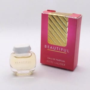 Peut inclure: Un petit flacon de parfum avec un bouchon blanc et un corps en verre transparent, à côté d'une boîte rose. Le flacon contient un liquide jaune clair et porte l'inscription "BEAUTIFUL". La boîte porte également l'inscription "BEAUTIFUL".