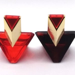 May include: Two triangular perfume bottles. One is bright red, the other is a deep burgundy. Both have gold-colored, triangular tops. The bottles are set against a white background.