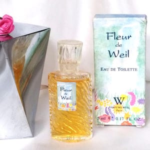 Può includere: Un piccolo flacone di profumo "Fleur de Weil" Eau de Toilette con la sua scatola abbinata. Il flacone è trasparente con un design strutturato e un tappo trasparente. La scatola è azzurra con decorazioni floreali e la scritta "Fleur de Weil". Contiene 5 ml.