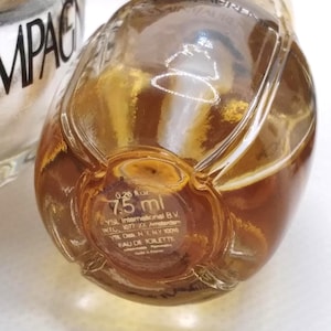 May include: Close-up of a small, clear glass perfume bottle with a golden-brown liquid inside. The bottle has a rounded shape and a gold-colored cap. The bottom of the bottle has text that reads "0.26 fl oz 7.5 ml".