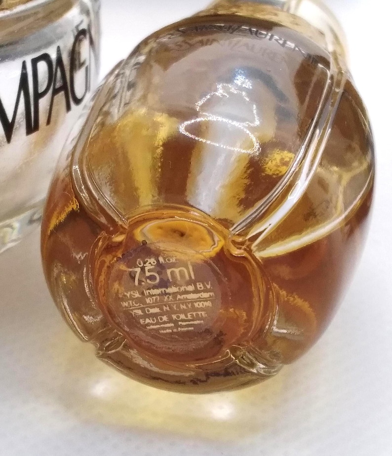 May include: Close-up of a small, clear glass perfume bottle with a golden-brown liquid inside. The bottle has a rounded shape and a gold-colored cap. The bottom of the bottle has text that reads "0.26 fl oz 7.5 ml".
