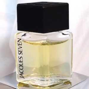 Pode incluir: Um frasco de perfume quadrado de vidro com tampa preta. O frasco contém um líquido amarelo e tem as palavras "JACQUES SEVEN" impressas verticalmente. O frasco está sobre uma superfície prateada reflexiva.