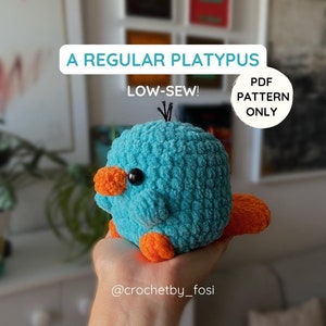 Op de afbeelding: Een gehaakt pluche platypus speelgoed met een blauw lichaam, een oranje snavel, voeten en staart. Het speelgoed wordt in een hand gehouden. De tekst "A REGULAR PLATYPUS LOW-SEW! PDF PATTERN ONLY @crochetby_fosi" is zichtbaar in de afbeelding.