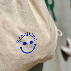 Könnte beinhalten: Beige Stoffbeutel mit Kordelzug, bedruckt mit dem Wort "Schulkind" und einem blauen Smiley-Gesicht. Der Beutel besteht aus hellem Stoff und hängt vor einem strukturierten, hellgrauen Hintergrund.
