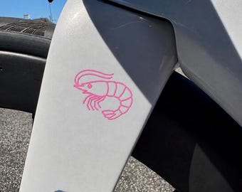 Könnte beinhalten: Eine hellgraue Fahrradgabel mit einer kleinen, rosafarbenen Strichzeichnung einer Garnele. Die Gabel ist über einem schwarzen Reifen positioniert, wobei ein Teil des schwarzen Lenkers oben im Rahmen vor einem klaren blauen Himmel zu sehen ist.