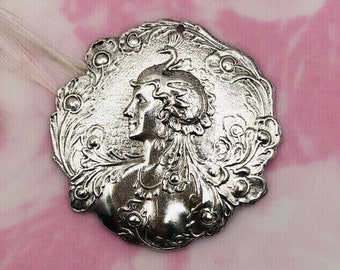 May include: Silver-toned metal pendant featuring an ornate design of a woman's profile with intricate floral details.