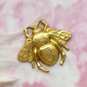 Könnte beinhalten: Ein goldfarbener Metall-Bienen-Anhänger mit detaillierten Flügeln und Beinen. Die Biene befindet sich auf einem rosa und weißen Hintergrund.