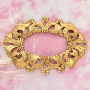 May include: A gold-toned metal frame with an ornate design, featuring intricate swirls and scrolls. The frame is oval-shaped and has a smooth, polished finish.