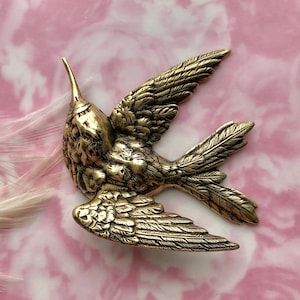 Pode incluir: Uma figurinha de beija-flor de metal dourado com asas detalhadas e um corpo texturizado. A figurinha está pousada sobre um fundo rosa e branco.