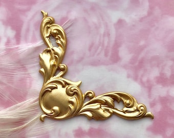May include: Gold-toned metal corner ornament with a decorative, ornate design. The ornament has a rounded center and intricate scrollwork.