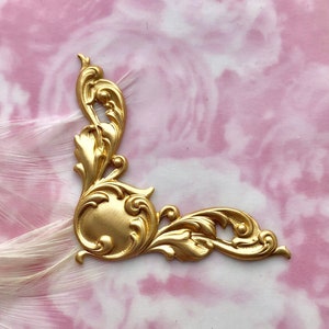 May include: Gold-toned metal corner ornament with a decorative, ornate design. The ornament has a rounded center and intricate scrollwork.