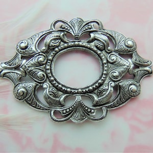 May include: Ornate silver-colored metal oval frame with a central circular opening. The frame features detailed scrollwork, floral motifs, and beaded accents. Ideal for jewelry making or crafting projects.