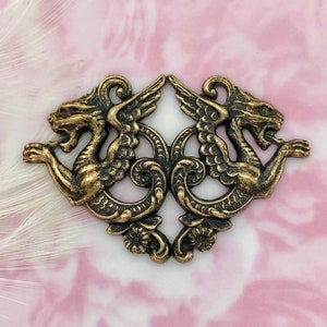 May include: A pair of gold-toned metal decorative embellishments featuring a winged griffin design. Each griffin is facing outward with its wings spread. The embellishments are connected by a decorative scrollwork design.
