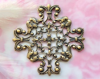 May include: Ornate, gold-colored filigree metalwork in a symmetrical cross shape. The design features scrolling vines, fleur-de-lis elements, and small beaded accents. The filigree is set against a soft pink and white background.