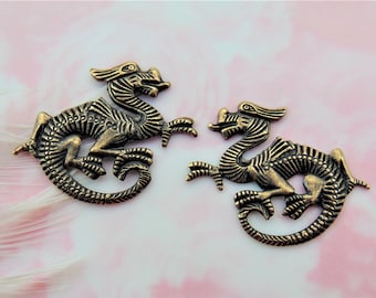 Op de afbeelding: Twee antieke goudkleurige metalen drakencharms. De draken zijn in een cirkelvormige vorm opgerold met hun staarten in hun mond. De draken hebben gedetailleerde schubben en vleugels.