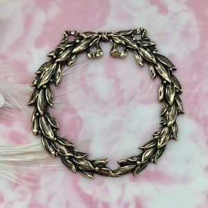 Pode incluir: Um amuleto de coroa de metal dourado com um design de folhas detalhado. A coroa é aberta na parte superior e inferior, permitindo que seja usado como um pingente ou enfeite.