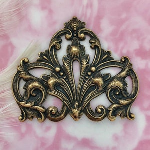 May include: Ornate bronze-colored metal embellishment with a symmetrical design. The decorative piece features scrolling flourishes, floral elements, and a central stylized leaf motif. The background is a soft pink and white marbled pattern.