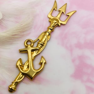 Puede incluir: Un amuleto de metal dorado con un tridente y un ancla entrelazados. El amuleto tiene un diseño detallado y un acabado pulido.