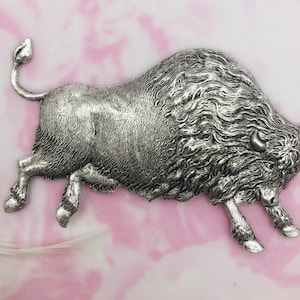 May include: A silver-colored metal bison figurine in a running pose. The bison has detailed fur and a curved tail. The background is a soft pink and white watercolor-like pattern.