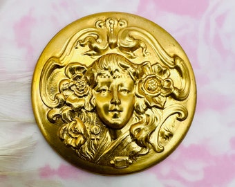 May include: A gold-toned metal brooch featuring a raised design of a woman's face surrounded by flowers. The brooch is circular and has a detailed, ornate design.