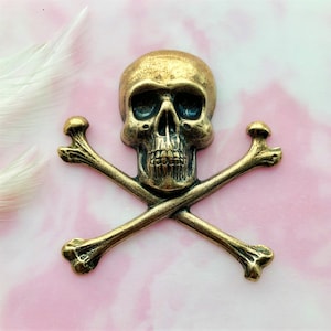 May include: An antique-style brass skull and crossbones emblem. The skull features hollow eye sockets and a detailed jaw. The crossed bones are positioned beneath the skull. The piece is set against a pink and white background.